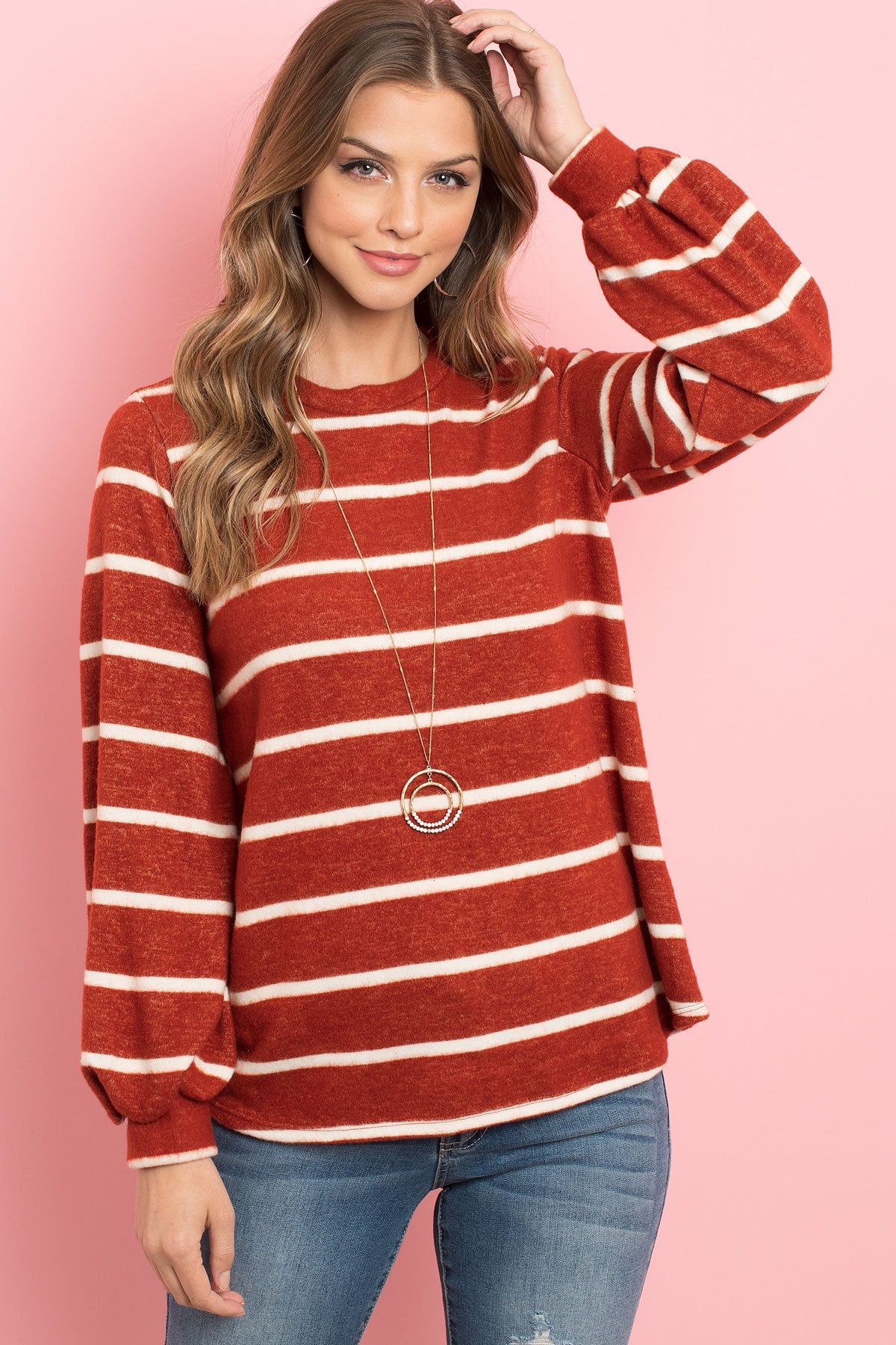 A woman in a Riah Fashion Striped Long Sleeved Round Neck Swing Top and jeans stands against a pink backdrop, wearing a long necklace and touching her hair.