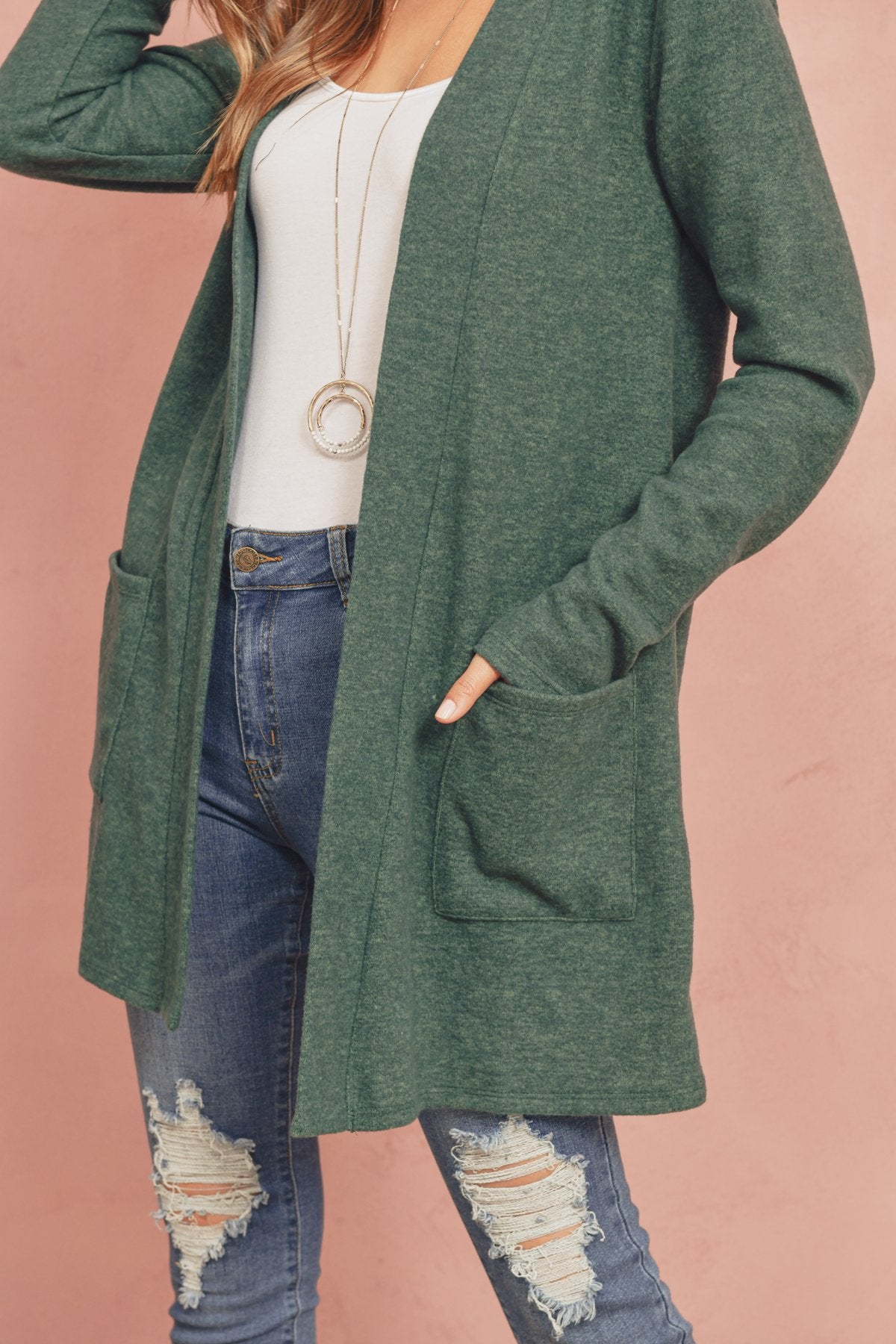 A woman in a Brushed Melange Open Front Cardigan by Riah Fashion and ripped jeans stands against a pink background, hands in her pockets.