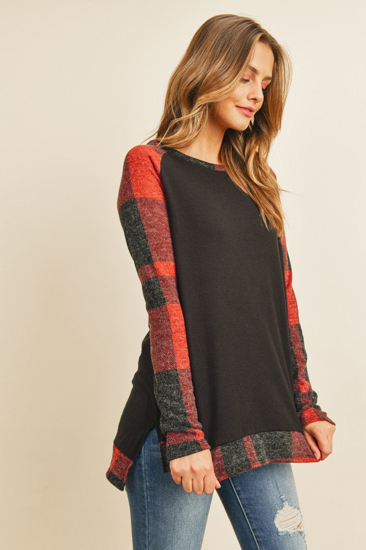 A woman in a Riah Fashion Brush Hacci Plaid Contrast Long Back Hem Top with red and beige accents, paired with blue ripped jeans, stands against a plain background.