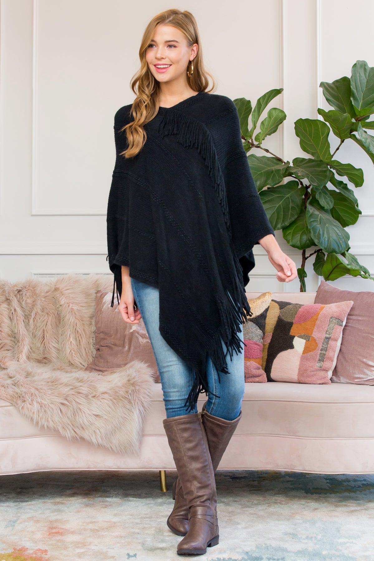 A person stands wearing a Riah Fashion Asymmetric Fringe Poncho in one color, paired with jeans and brown boots, smiling while holding the poncho. In the background are a plant and a sofa with cushions.