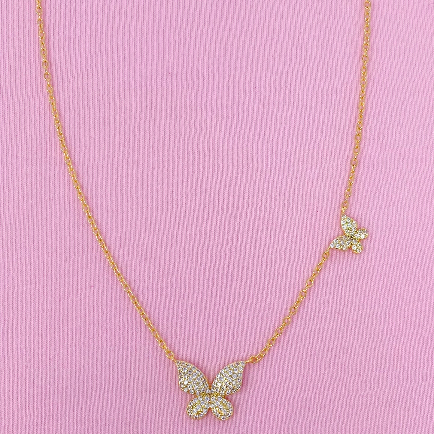 The Ellison + Young Double Shine Butterfly Necklace, featuring butterfly-shaped pendants adorned with small crystals, is elegantly displayed on a light marble background.