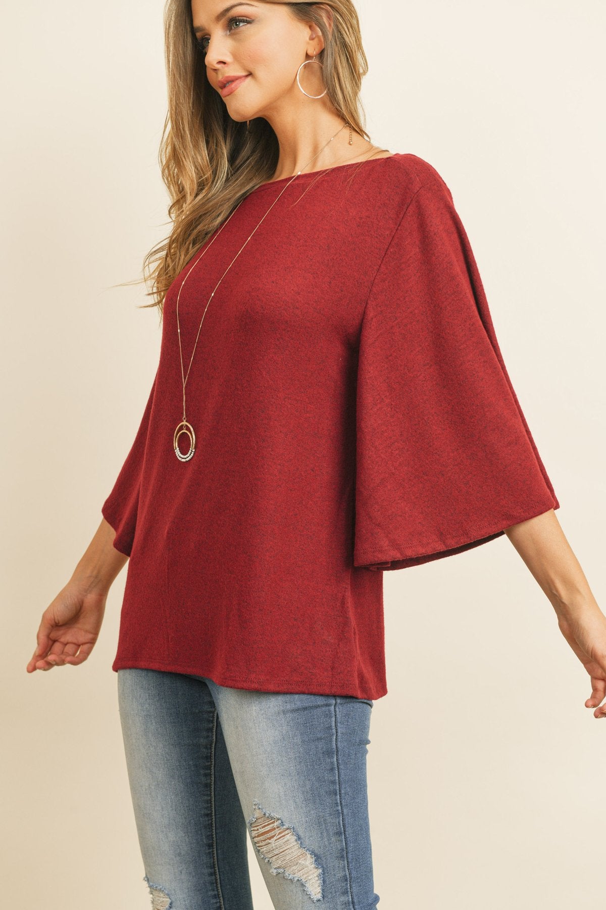 A person in a Riah Fashion Boat Neck Bell Sleeve Solid Hacci Brushed Top and ripped jeans stands against a neutral background, accessorized with hoop earrings and a long necklace.