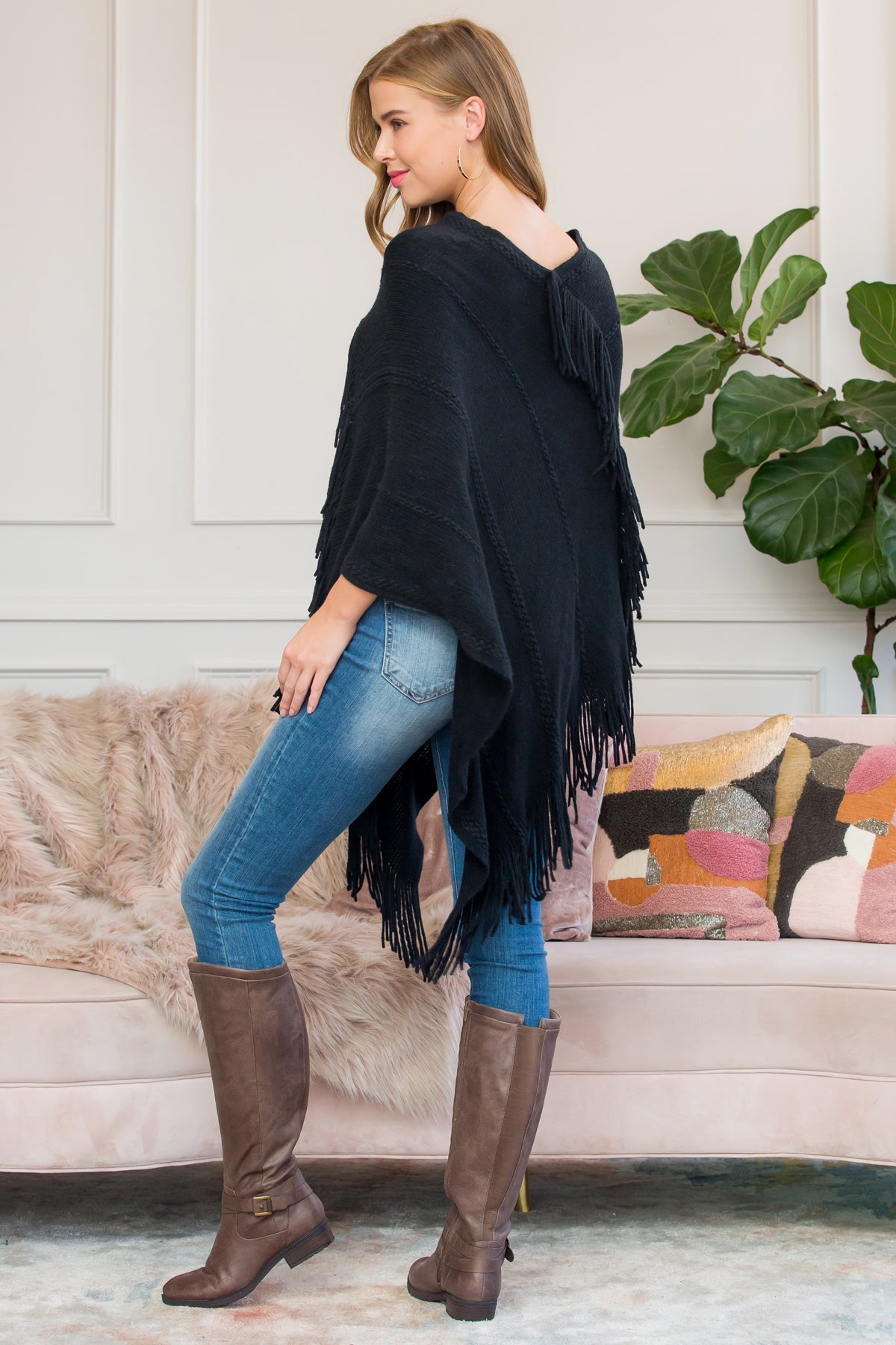 A person stands wearing a Riah Fashion Asymmetric Fringe Poncho in one color, paired with jeans and brown boots, smiling while holding the poncho. In the background are a plant and a sofa with cushions.
