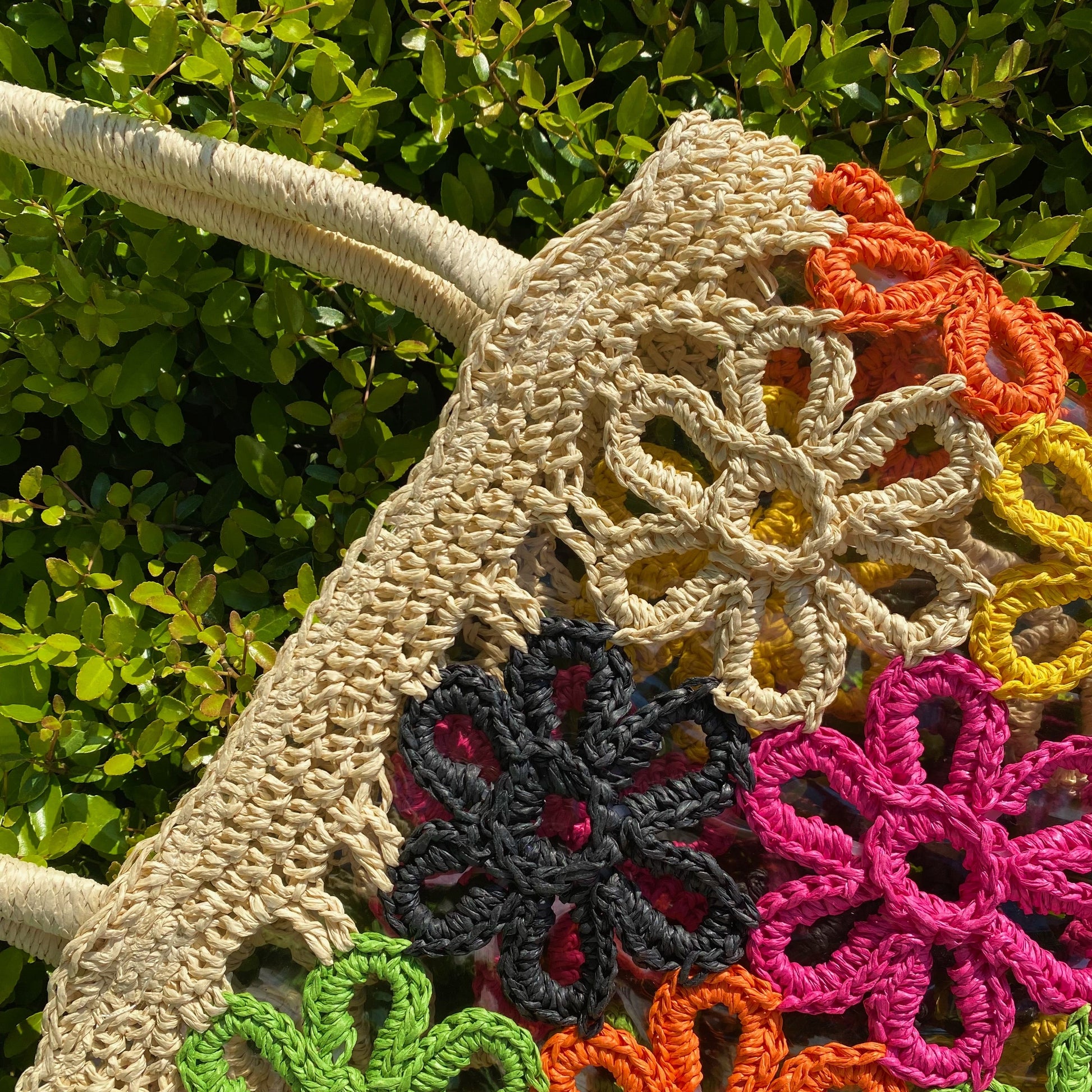 The Hand Made Flower Power Tote by Ellison + Young, featuring colorful floral crochet patterns in orange, green, pink, black, and yellow, rests on dense green foliage.