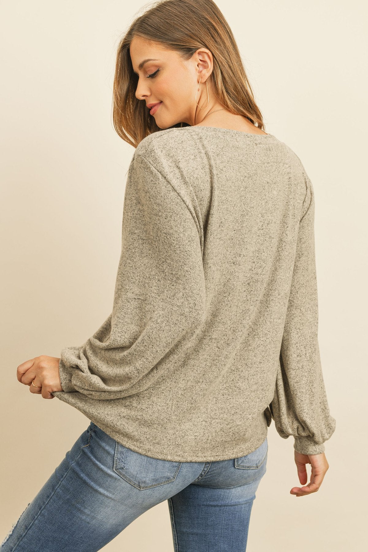 A woman models a Riah Fashion Puff Sleeved Boat Neck Two Toned Brushed Hacci Top with ripped jeans and brown boots on a concrete floor against a plain backdrop, accessorized with a long necklace.