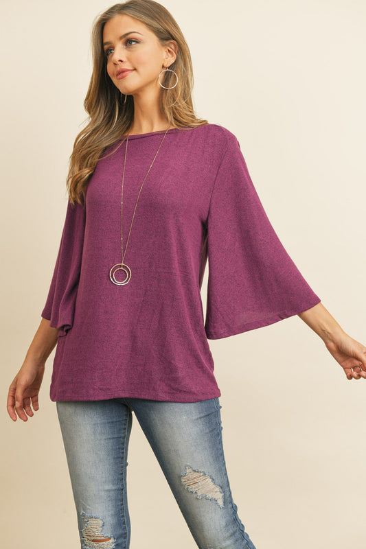 A person in a Riah Fashion Boat Neck Bell Sleeve Solid Hacci Brushed Top and ripped jeans stands against a neutral background, accessorized with hoop earrings and a long necklace.