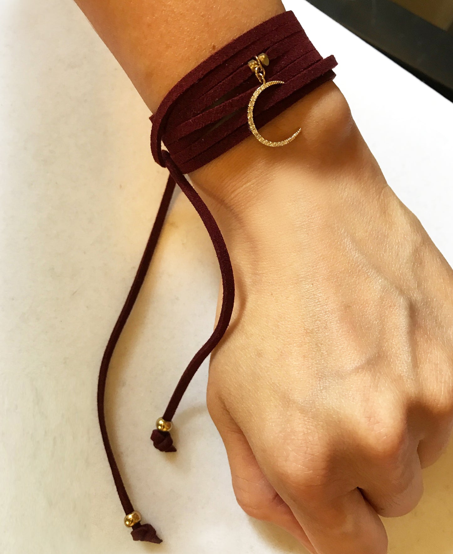 The Crescent Moon Burgundy Suede Wrap Bracelet by Gena Myint is a dark red suede bracelet featuring a gold crescent moon charm adorned with small crystals.