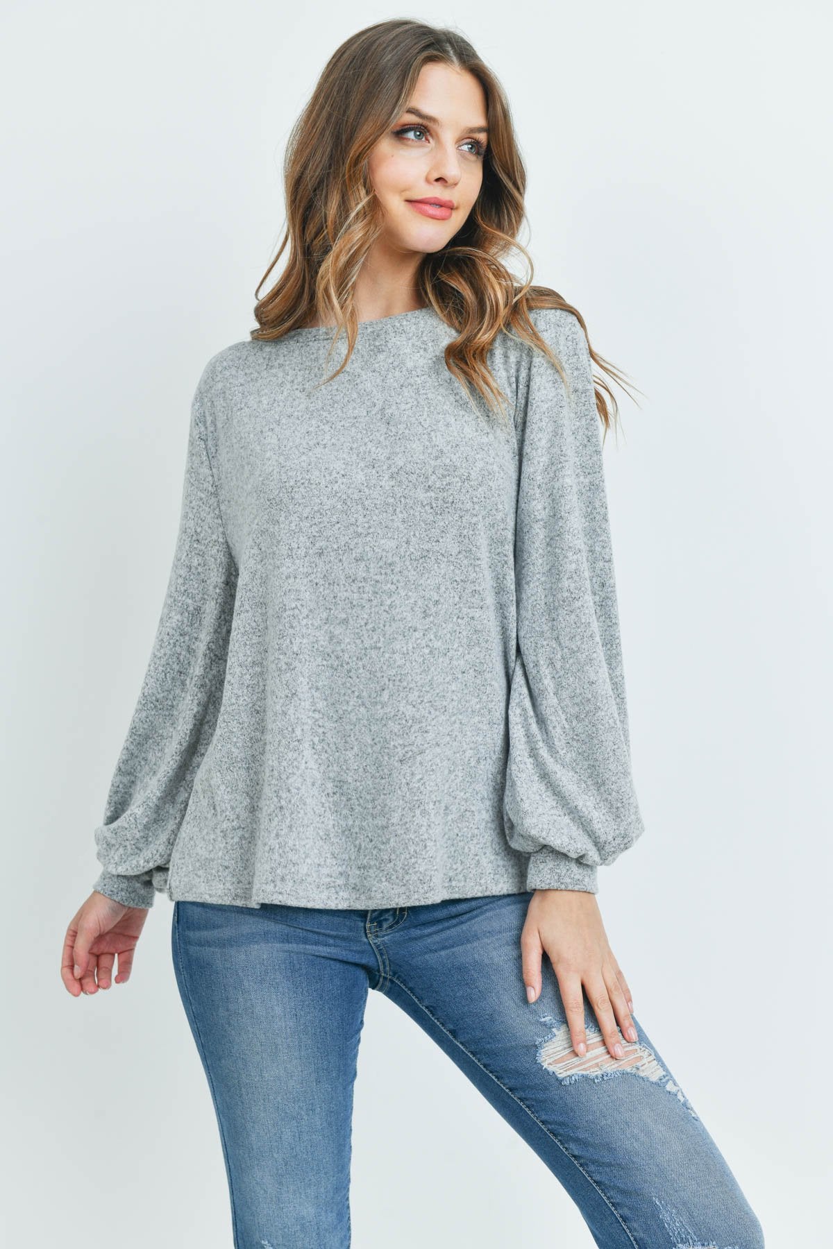 A woman models a Riah Fashion Puff Sleeved Boat Neck Two Toned Brushed Hacci Top with ripped jeans and brown boots on a concrete floor against a plain backdrop, accessorized with a long necklace.