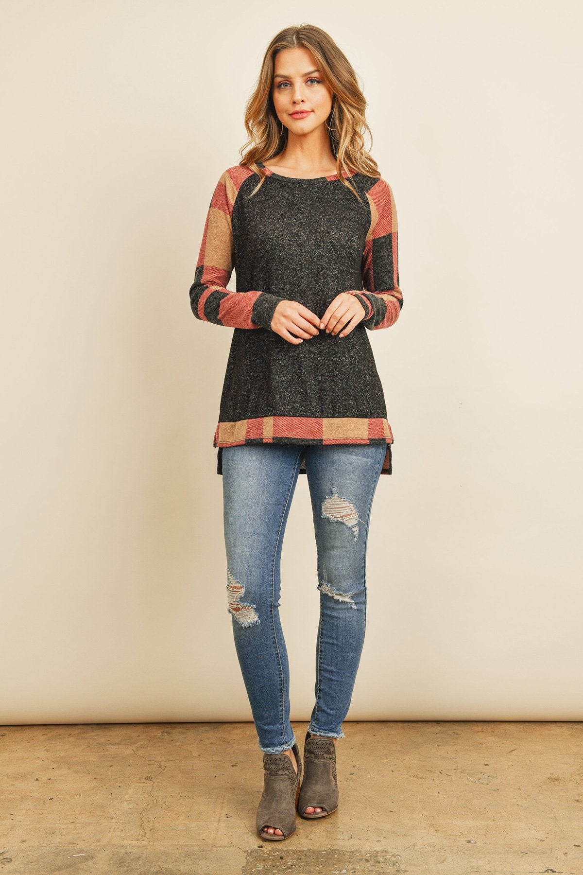 A woman in a Riah Fashion Brush Hacci Plaid Contrast Long Back Hem Top with red and beige accents, paired with blue ripped jeans, stands against a plain background.