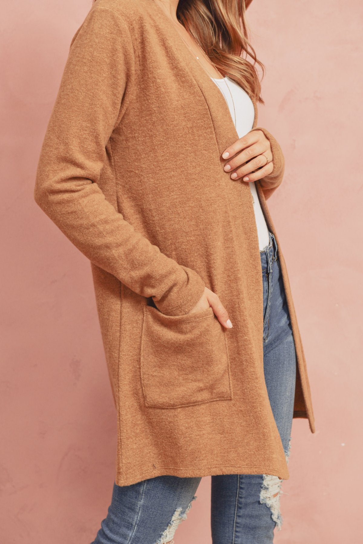 A woman in a Brushed Melange Open Front Cardigan by Riah Fashion and ripped jeans stands against a pink background, hands in her pockets.