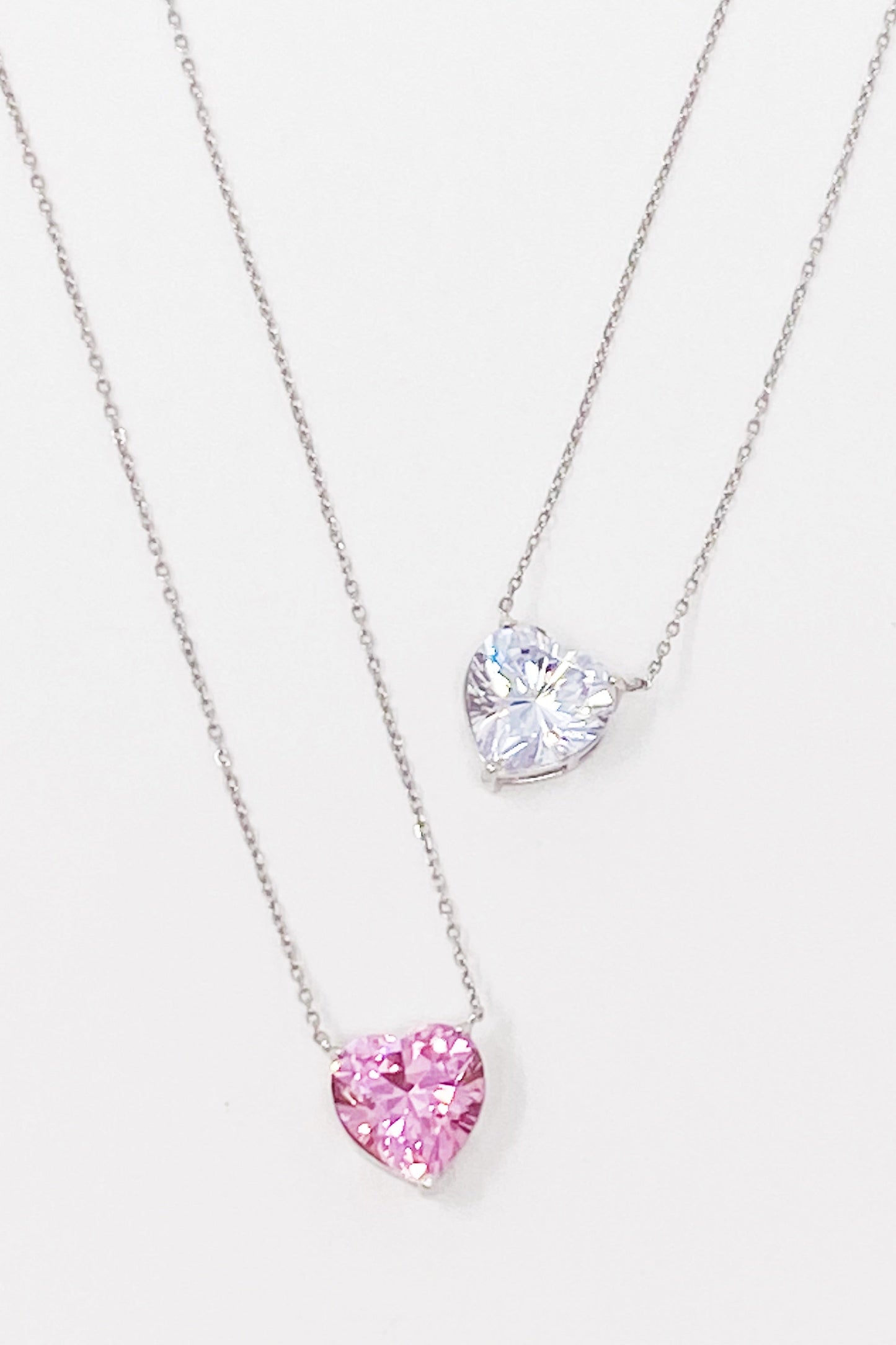 Two Absolute Beauty sterling silver heart necklaces by Ellison + Young; one with a pink gem, the other with a clear gem, against a white background.