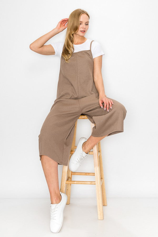 A person with long blonde hair sits on a wooden stool against a plain background, wearing Stylespects Cropped Bottom Wide Leg Oversized Jumpsuit in taupe and white sneakers.