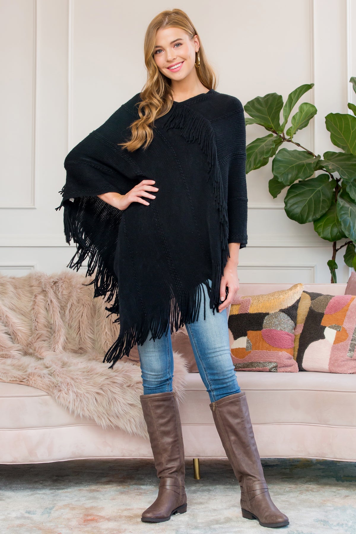 A person stands wearing a Riah Fashion Asymmetric Fringe Poncho in one color, paired with jeans and brown boots, smiling while holding the poncho. In the background are a plant and a sofa with cushions.