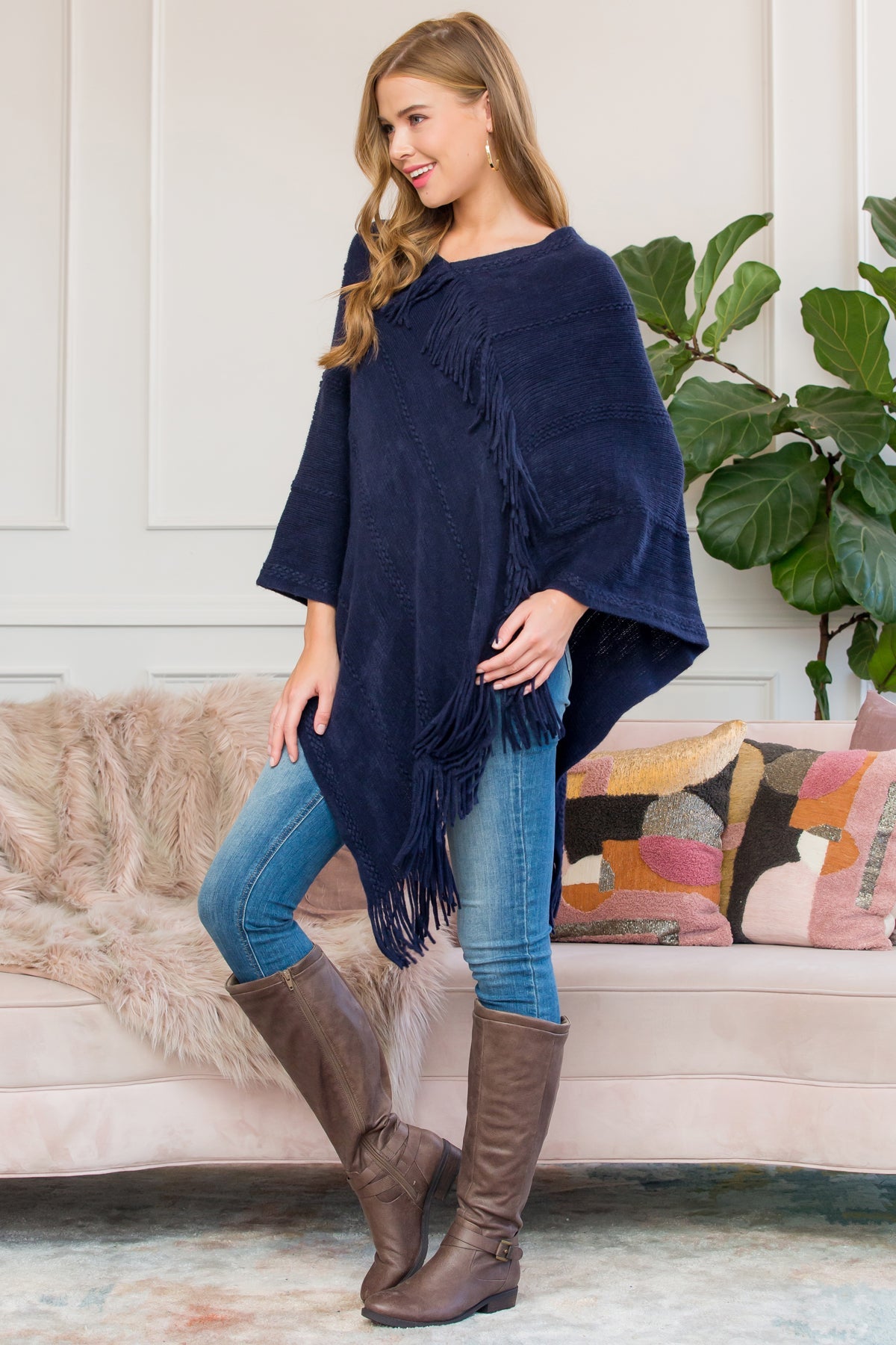 A person stands wearing a Riah Fashion Asymmetric Fringe Poncho in one color, paired with jeans and brown boots, smiling while holding the poncho. In the background are a plant and a sofa with cushions.