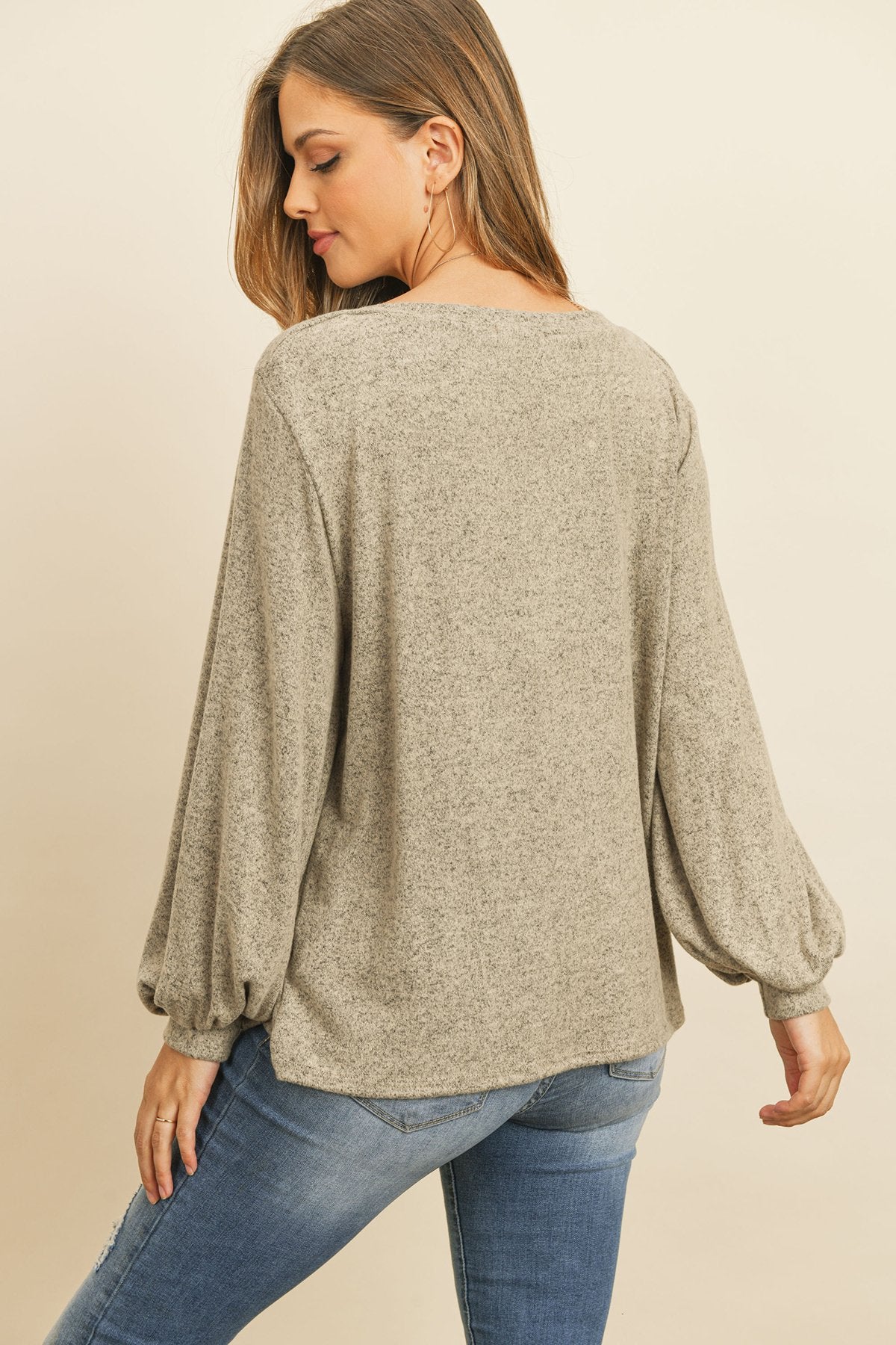 A woman models a Riah Fashion Puff Sleeved Boat Neck Two Toned Brushed Hacci Top with ripped jeans and brown boots on a concrete floor against a plain backdrop, accessorized with a long necklace.