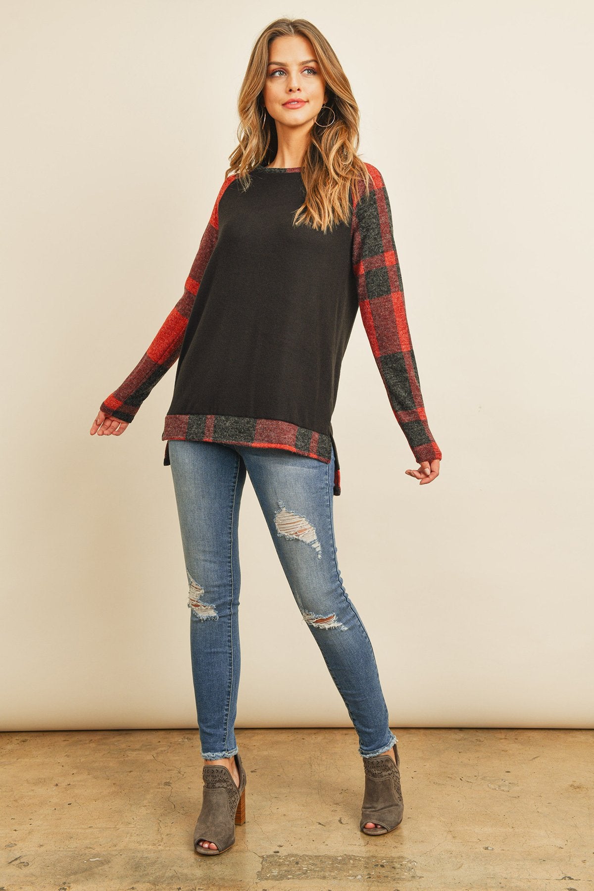 A woman in a Riah Fashion Brush Hacci Plaid Contrast Long Back Hem Top with red and beige accents, paired with blue ripped jeans, stands against a plain background.