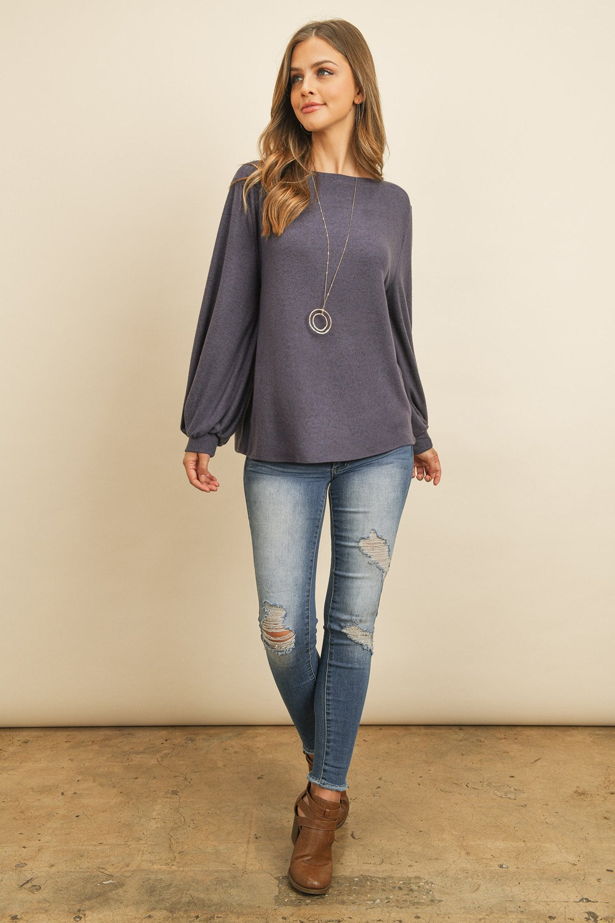 A woman models a Riah Fashion Puff Sleeved Boat Neck Two Toned Brushed Hacci Top with ripped jeans and brown boots on a concrete floor against a plain backdrop, accessorized with a long necklace.