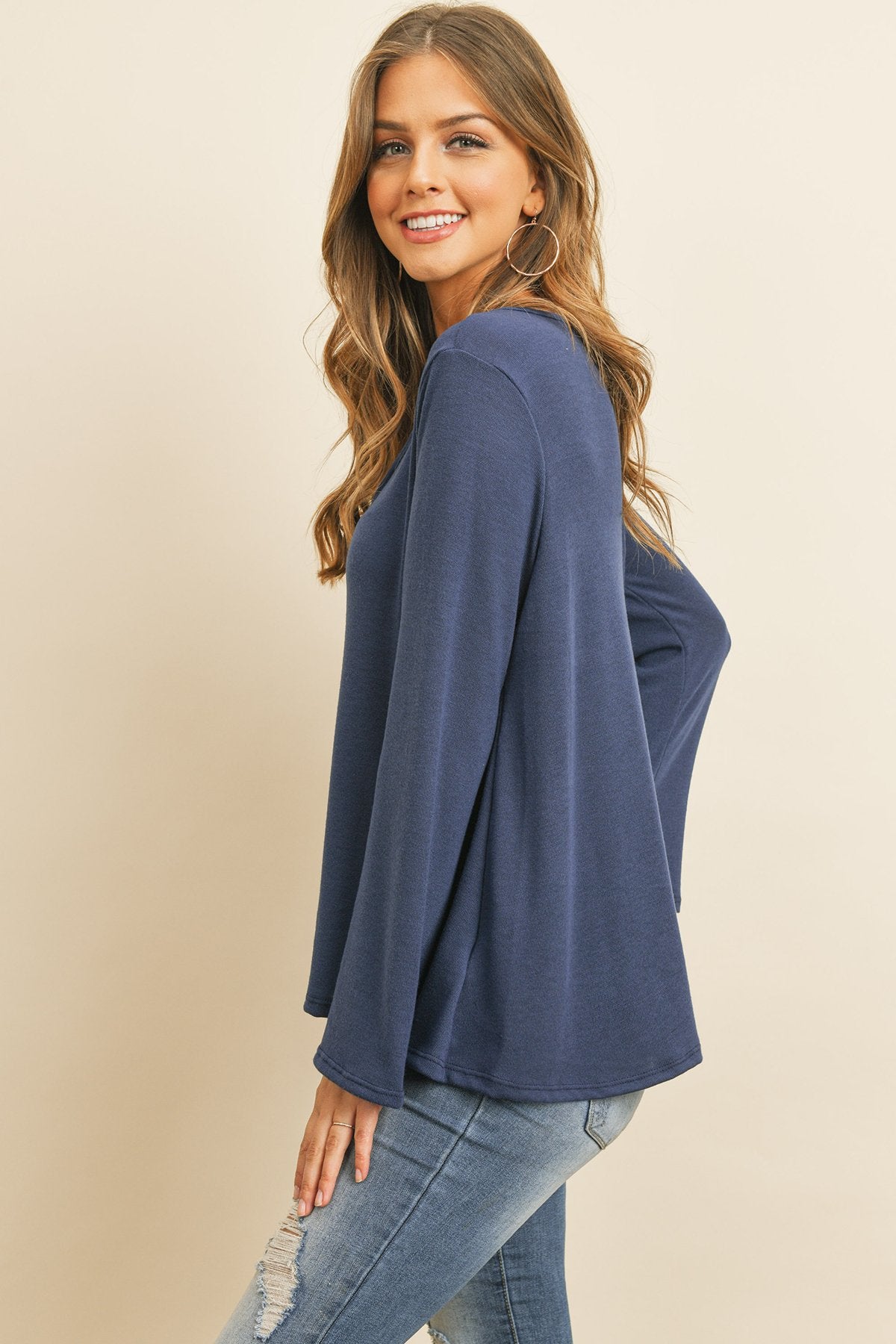 A person wearing a Riah Fashion Criss Cross Neck Long Sleeve Hacci Top in Dusty Blush and blue jeans stands against a neutral background, smiling.