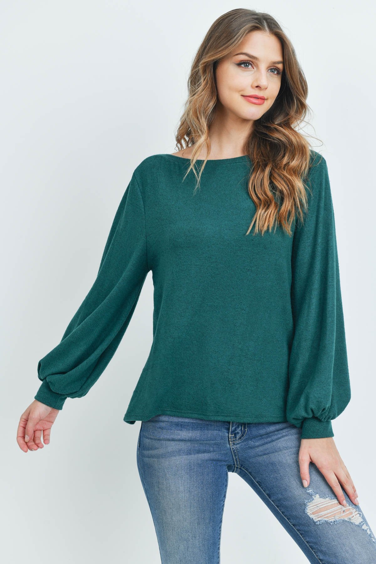 A woman models a Riah Fashion Puff Sleeved Boat Neck Two Toned Brushed Hacci Top with ripped jeans and brown boots on a concrete floor against a plain backdrop, accessorized with a long necklace.