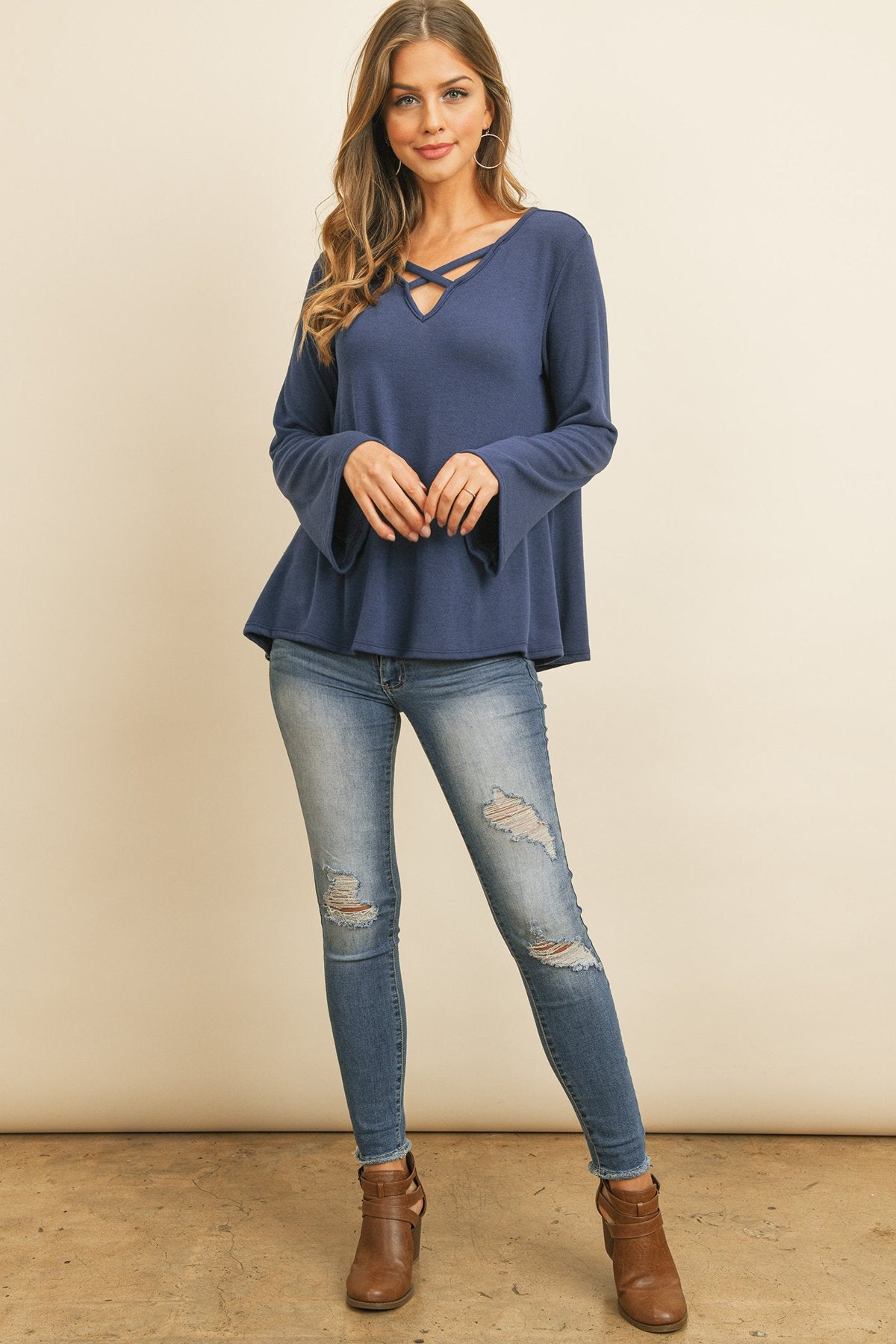 A person wearing a Riah Fashion Criss Cross Neck Long Sleeve Hacci Top in Dusty Blush and blue jeans stands against a neutral background, smiling.