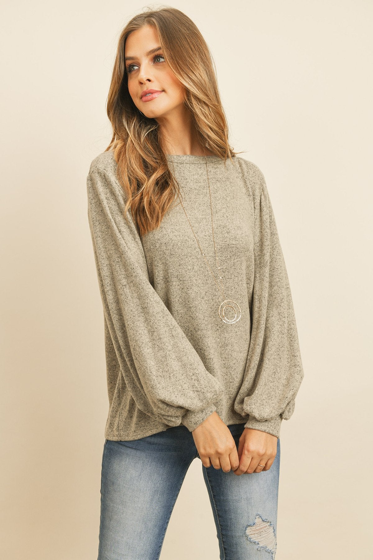 A woman models a Riah Fashion Puff Sleeved Boat Neck Two Toned Brushed Hacci Top with ripped jeans and brown boots on a concrete floor against a plain backdrop, accessorized with a long necklace.