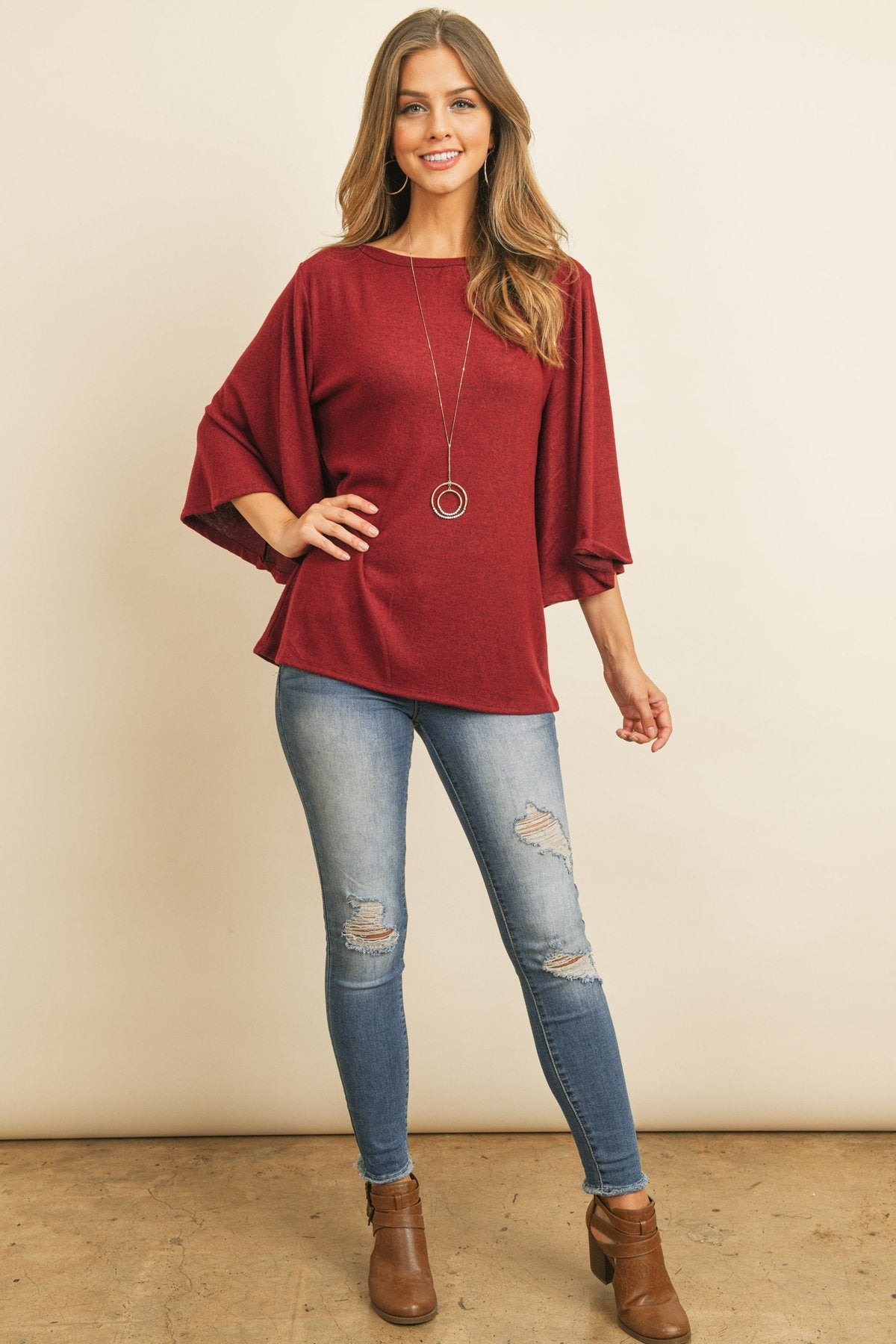 A person in a Riah Fashion Boat Neck Bell Sleeve Solid Hacci Brushed Top and ripped jeans stands against a neutral background, accessorized with hoop earrings and a long necklace.