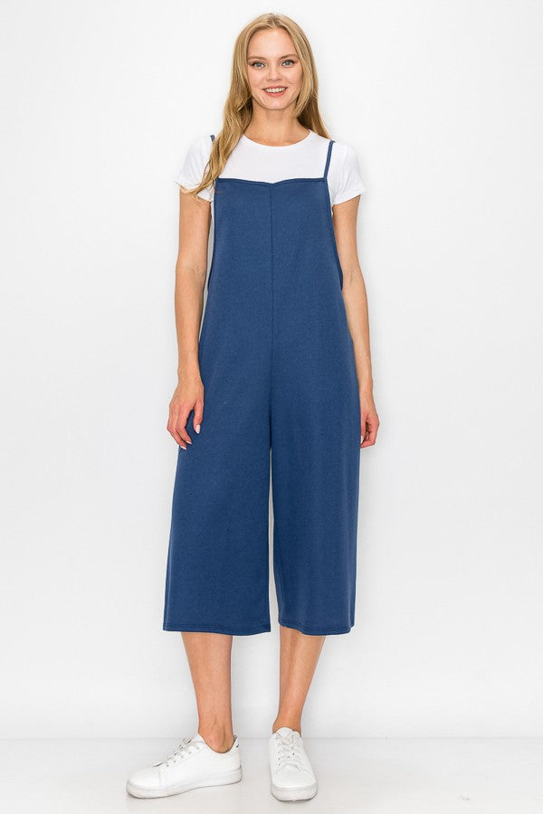 A person wearing a Stylespect Cropped Bottom Wide Leg Oversized Jumpsuit in blue over a white T-shirt and white sneakers, standing and smiling against a plain white background.