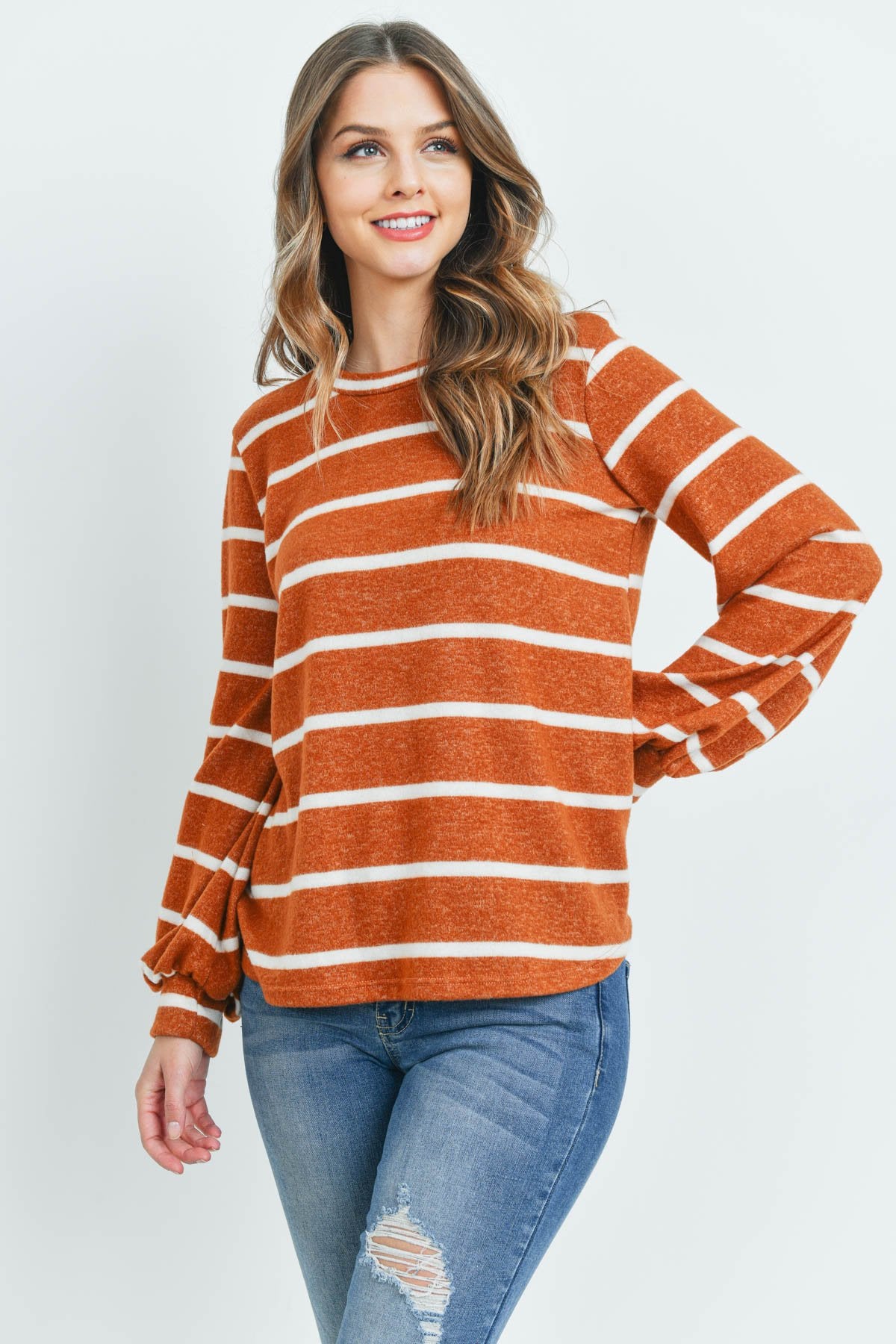 A woman in a Riah Fashion Striped Long Sleeved Round Neck Swing Top and jeans stands against a pink backdrop, wearing a long necklace and touching her hair.