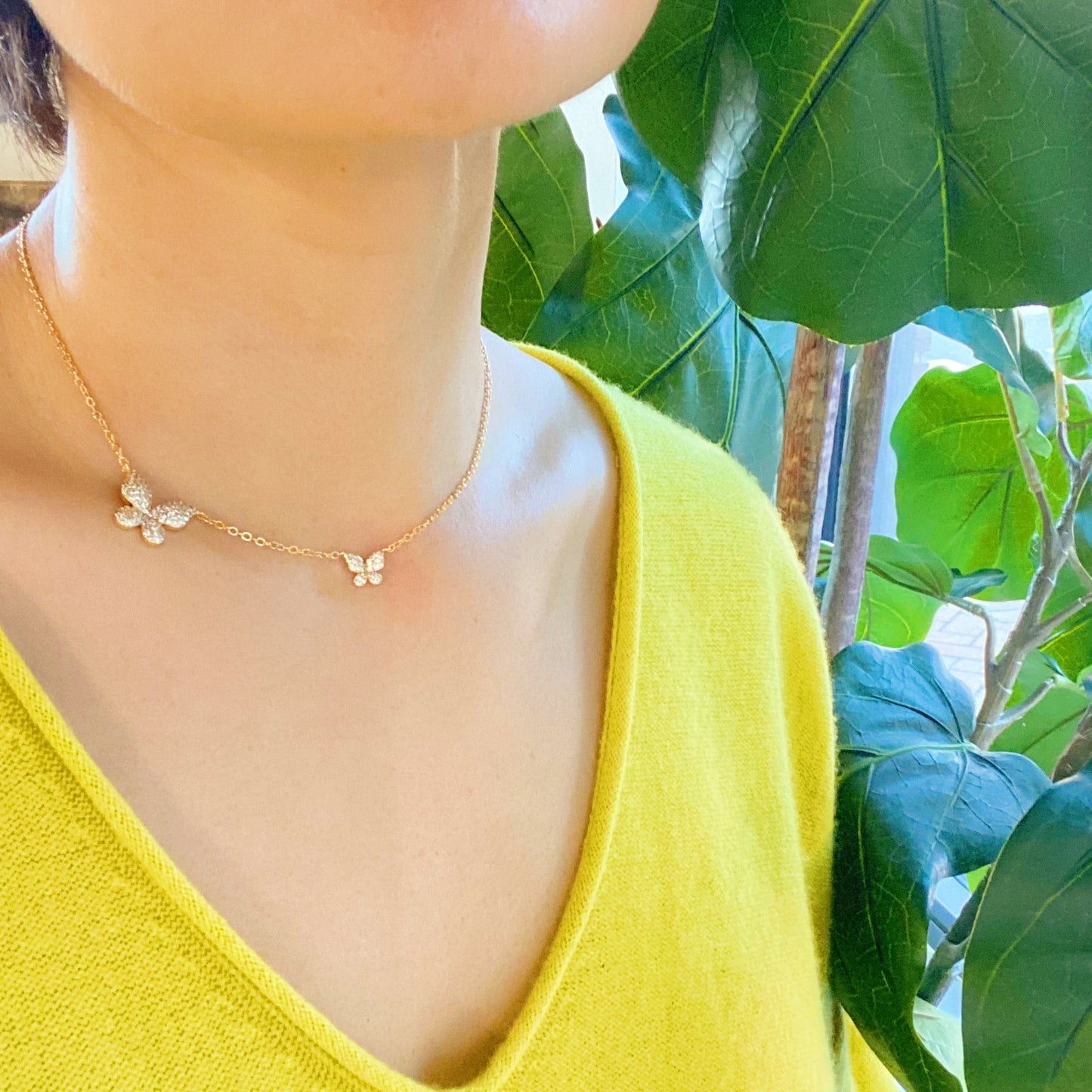 The Ellison + Young Double Shine Butterfly Necklace, featuring butterfly-shaped pendants adorned with small crystals, is elegantly displayed on a light marble background.