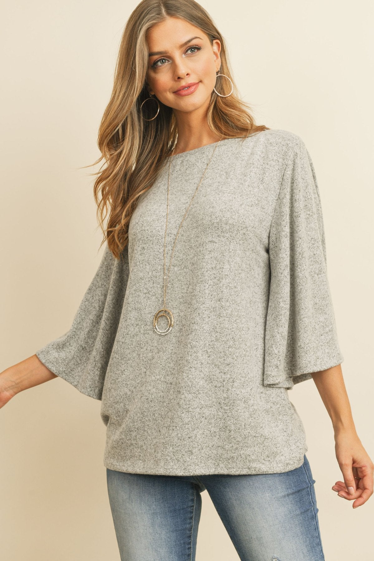 A person in a Riah Fashion Boat Neck Bell Sleeve Solid Hacci Brushed Top and ripped jeans stands against a neutral background, accessorized with hoop earrings and a long necklace.