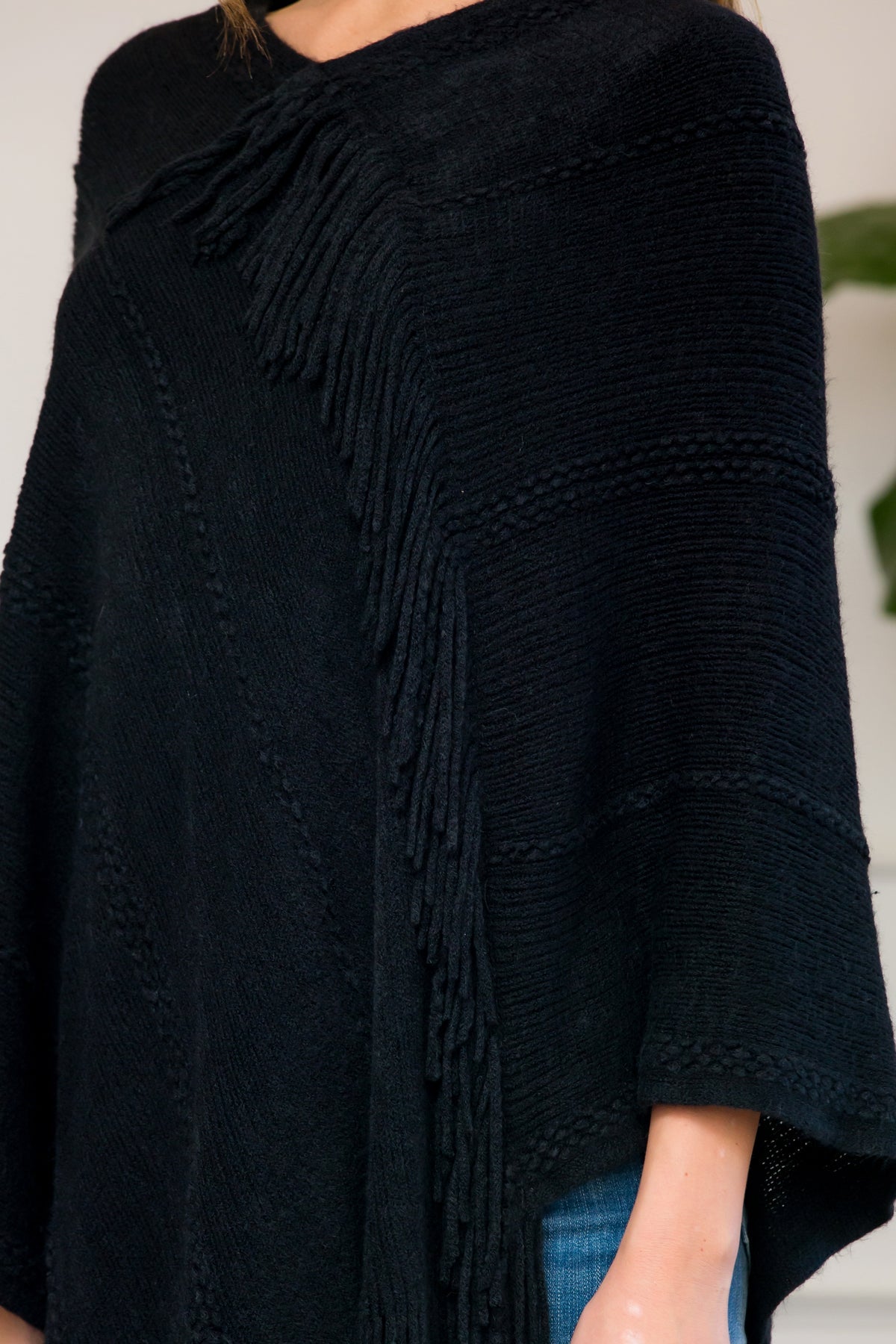 A person stands wearing a Riah Fashion Asymmetric Fringe Poncho in one color, paired with jeans and brown boots, smiling while holding the poncho. In the background are a plant and a sofa with cushions.