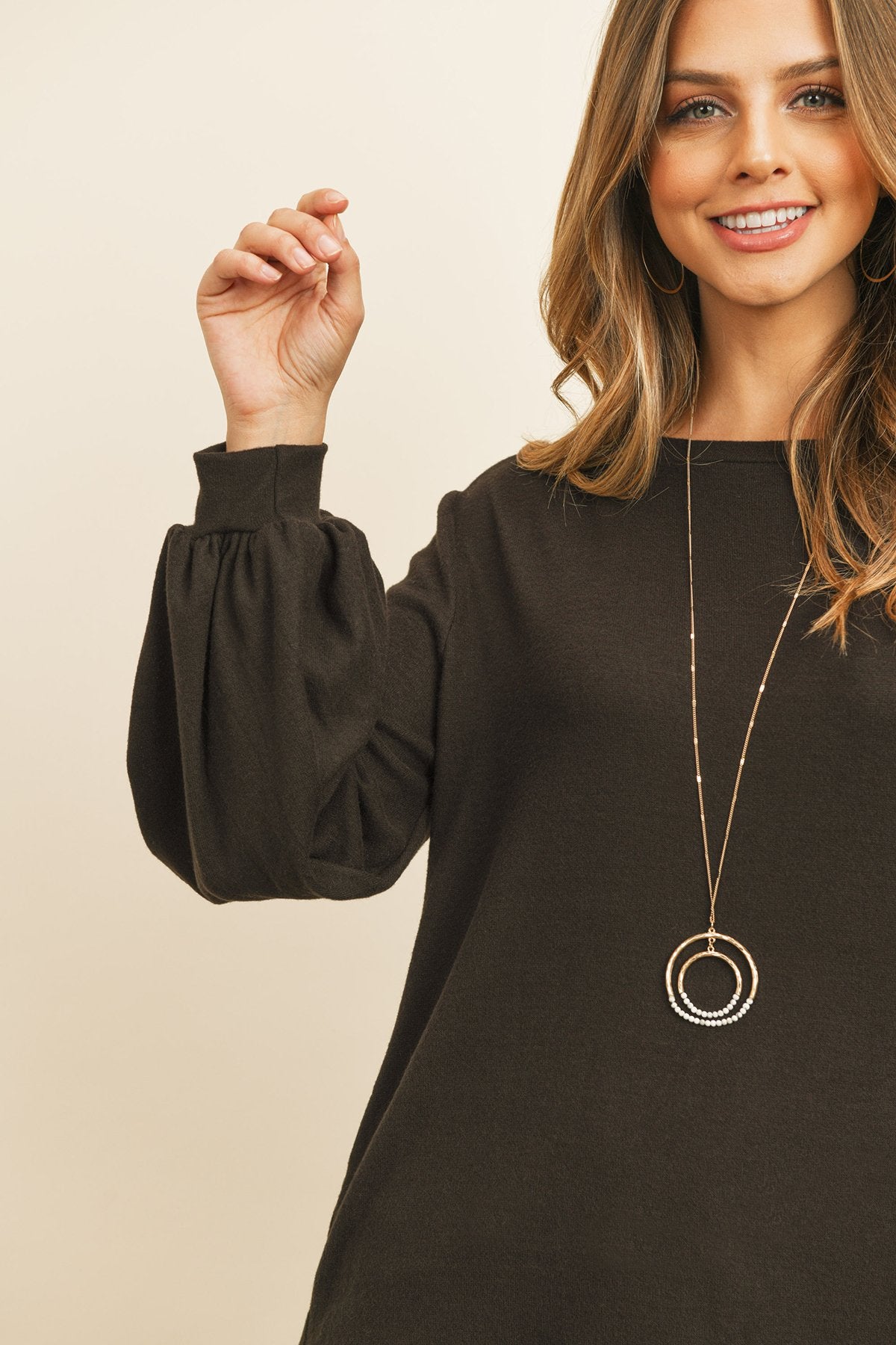 A woman models a Riah Fashion Puff Sleeved Boat Neck Two Toned Brushed Hacci Top with ripped jeans and brown boots on a concrete floor against a plain backdrop, accessorized with a long necklace.