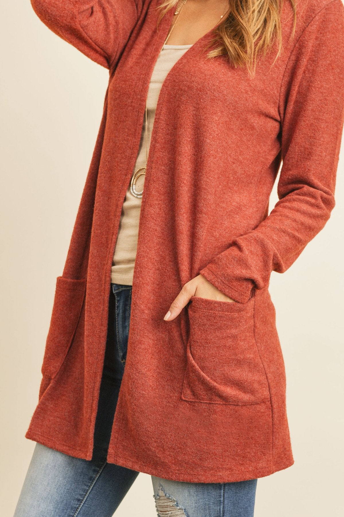 A woman in a teal Solid Long Sleeved Open Front Pocket Cardigan by Riah Fashion over a beige top stands against a pink background, smiling with hands in pockets.