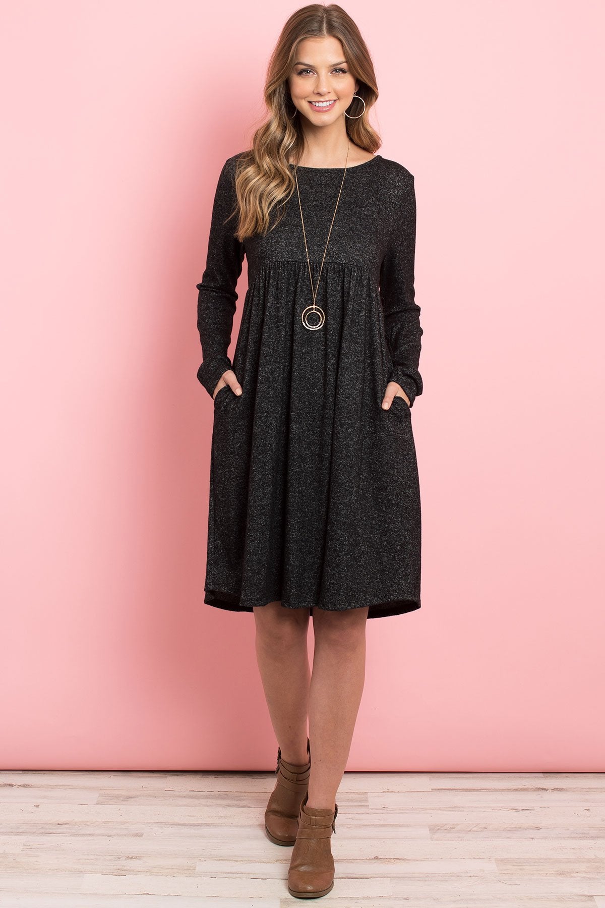A woman in a Riah Fashion Boat Neck Brushed Hacci Round Hem Dress in Dark Ruby stands before a pink backdrop, adorned with a long necklace featuring circular pendants.