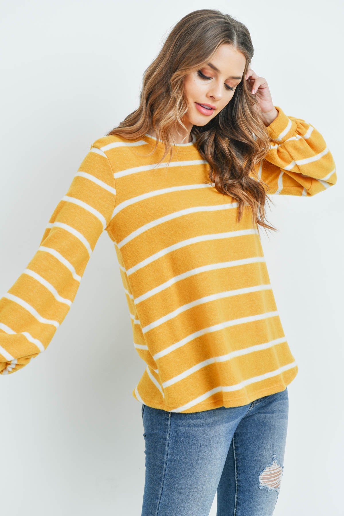 A woman in a Riah Fashion Striped Long Sleeved Round Neck Swing Top and jeans stands against a pink backdrop, wearing a long necklace and touching her hair.