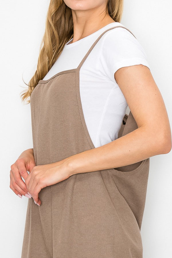 A person with long blonde hair sits on a wooden stool against a plain background, wearing Stylespects Cropped Bottom Wide Leg Oversized Jumpsuit in taupe and white sneakers.