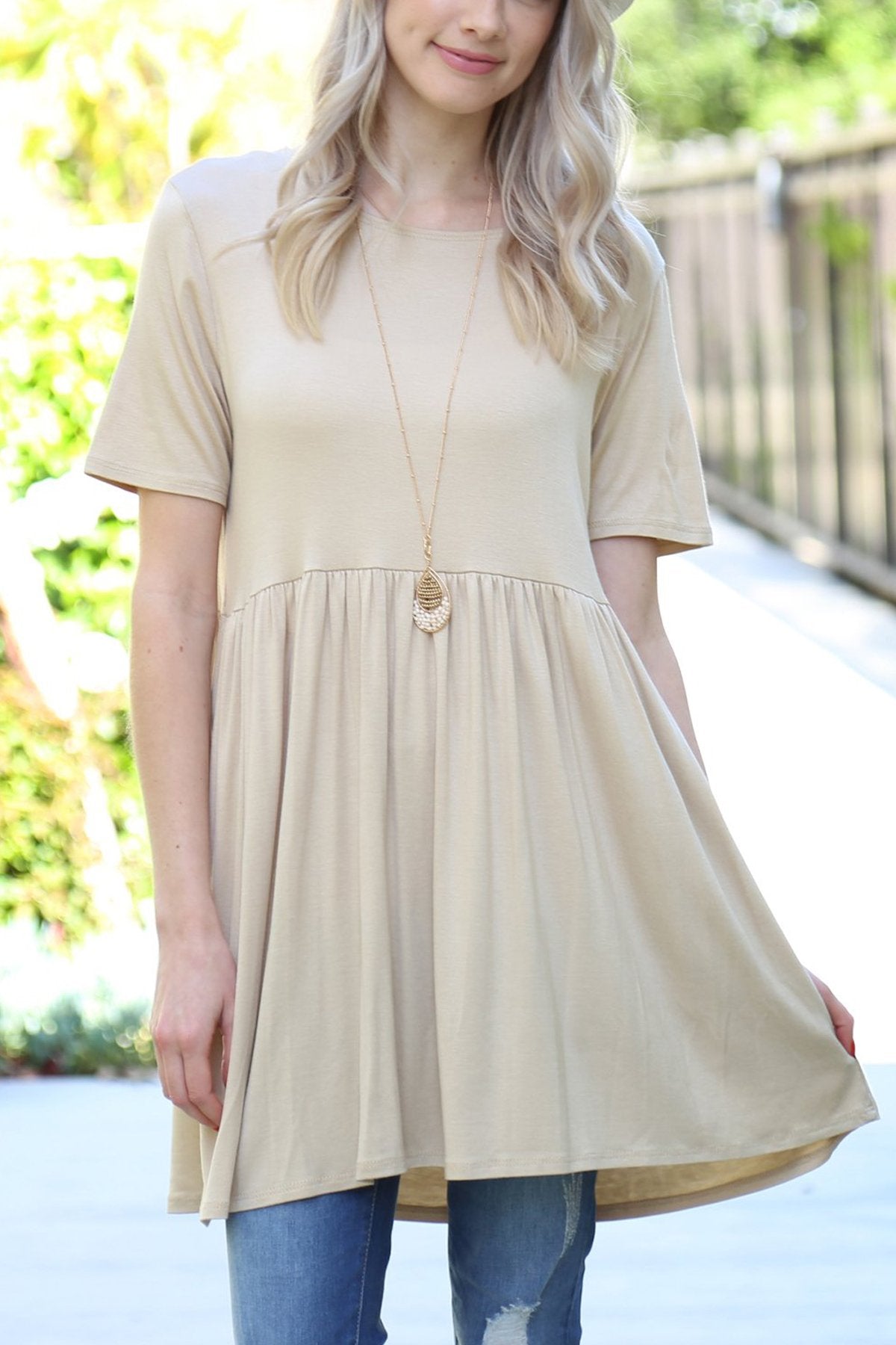 A woman stands outdoors wearing a beige Riah Fashion Short Sleeve Empire Waist Tunic paired with a long necklace.