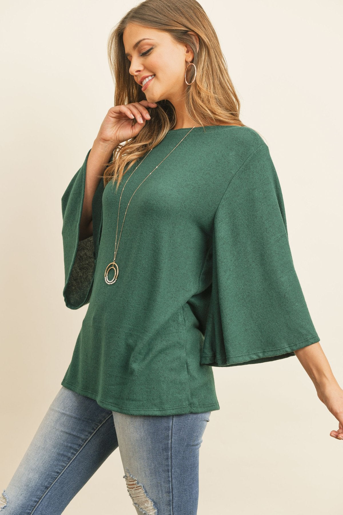 A person in a Riah Fashion Boat Neck Bell Sleeve Solid Hacci Brushed Top and ripped jeans stands against a neutral background, accessorized with hoop earrings and a long necklace.