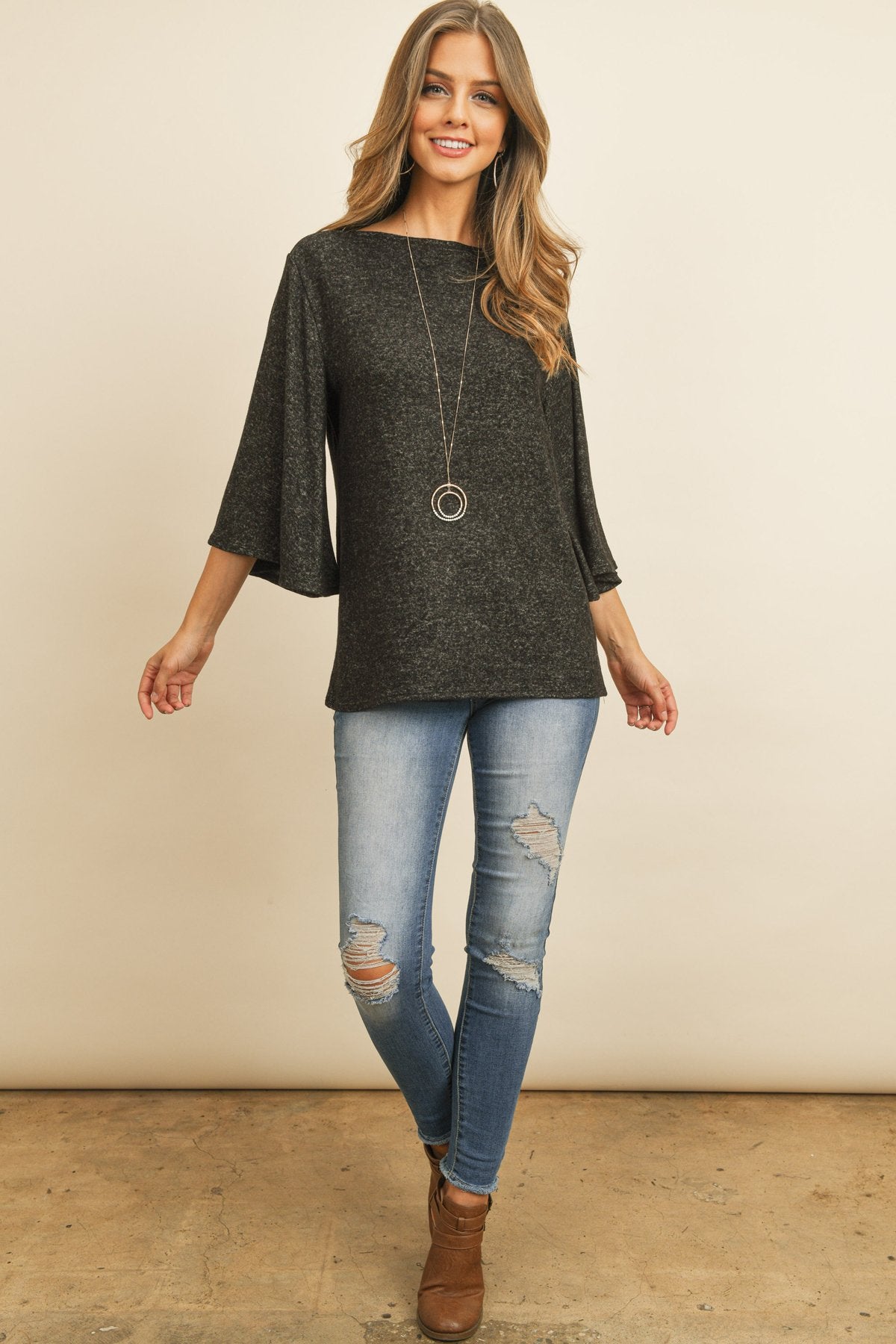 A person in a Riah Fashion Boat Neck Bell Sleeve Solid Hacci Brushed Top and ripped jeans stands against a neutral background, accessorized with hoop earrings and a long necklace.