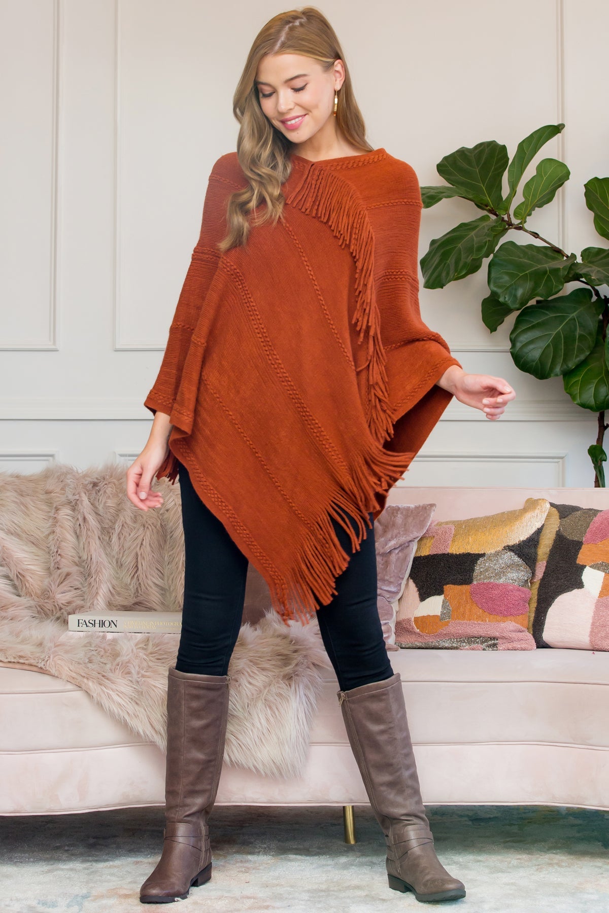 A person stands wearing a Riah Fashion Asymmetric Fringe Poncho in one color, paired with jeans and brown boots, smiling while holding the poncho. In the background are a plant and a sofa with cushions.
