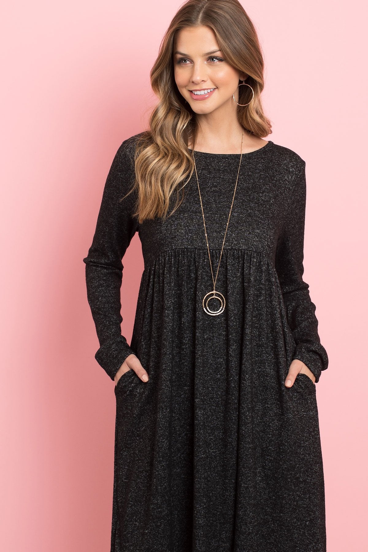 A woman in a Riah Fashion Boat Neck Brushed Hacci Round Hem Dress in Dark Ruby stands before a pink backdrop, adorned with a long necklace featuring circular pendants.