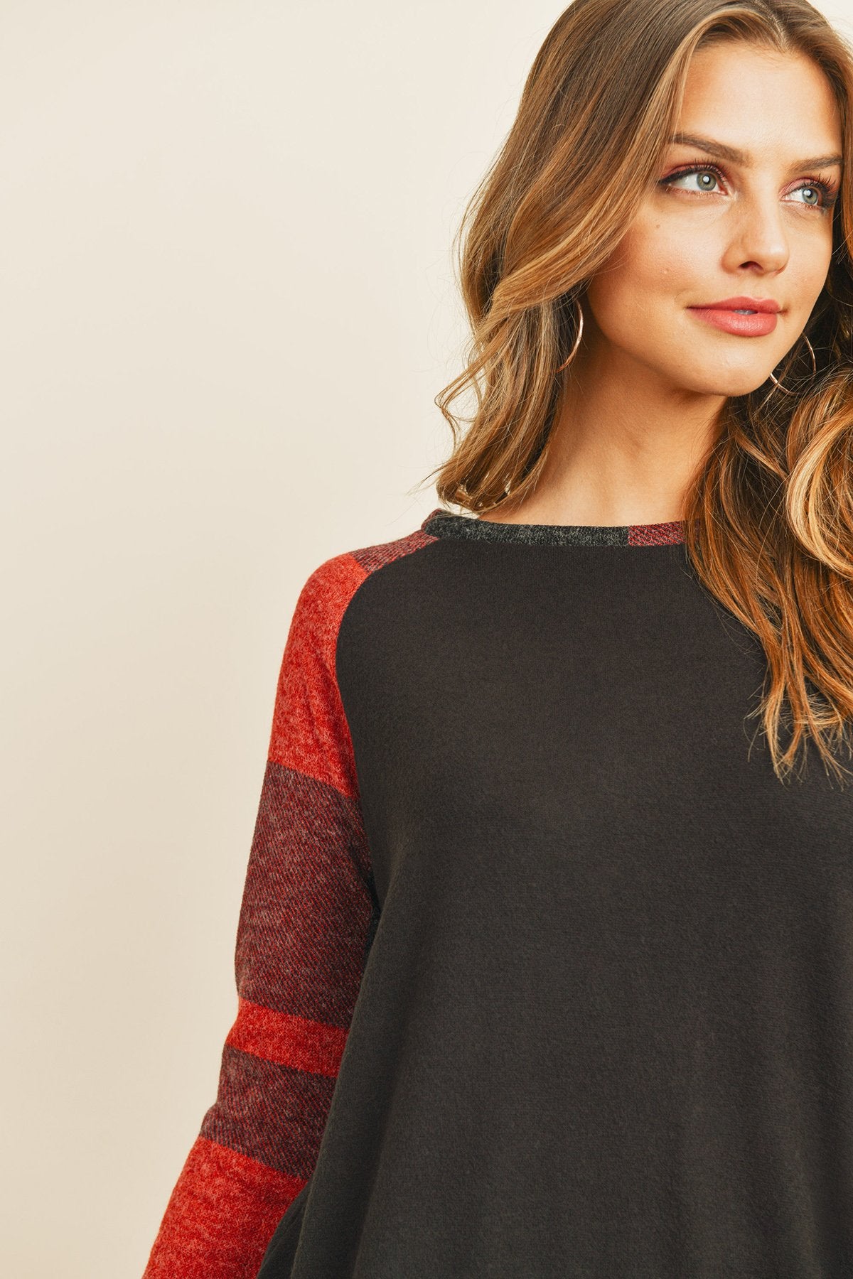 A woman in a Riah Fashion Brush Hacci Plaid Contrast Long Back Hem Top with red and beige accents, paired with blue ripped jeans, stands against a plain background.