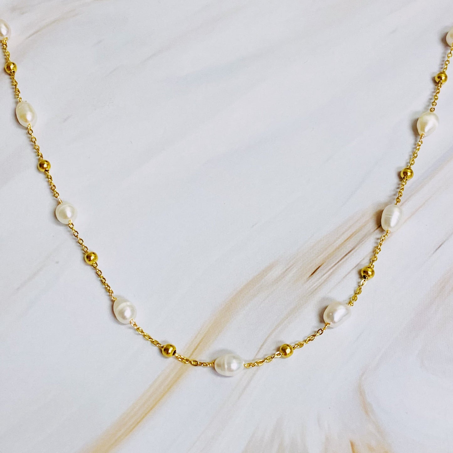 The Ashley Freshwater Pearl Necklace by Ellison + Young is a delicate gold chain adorned with small pearls and tiny gold beads, elegantly displayed on a light marble surface with green leaves and white stones.