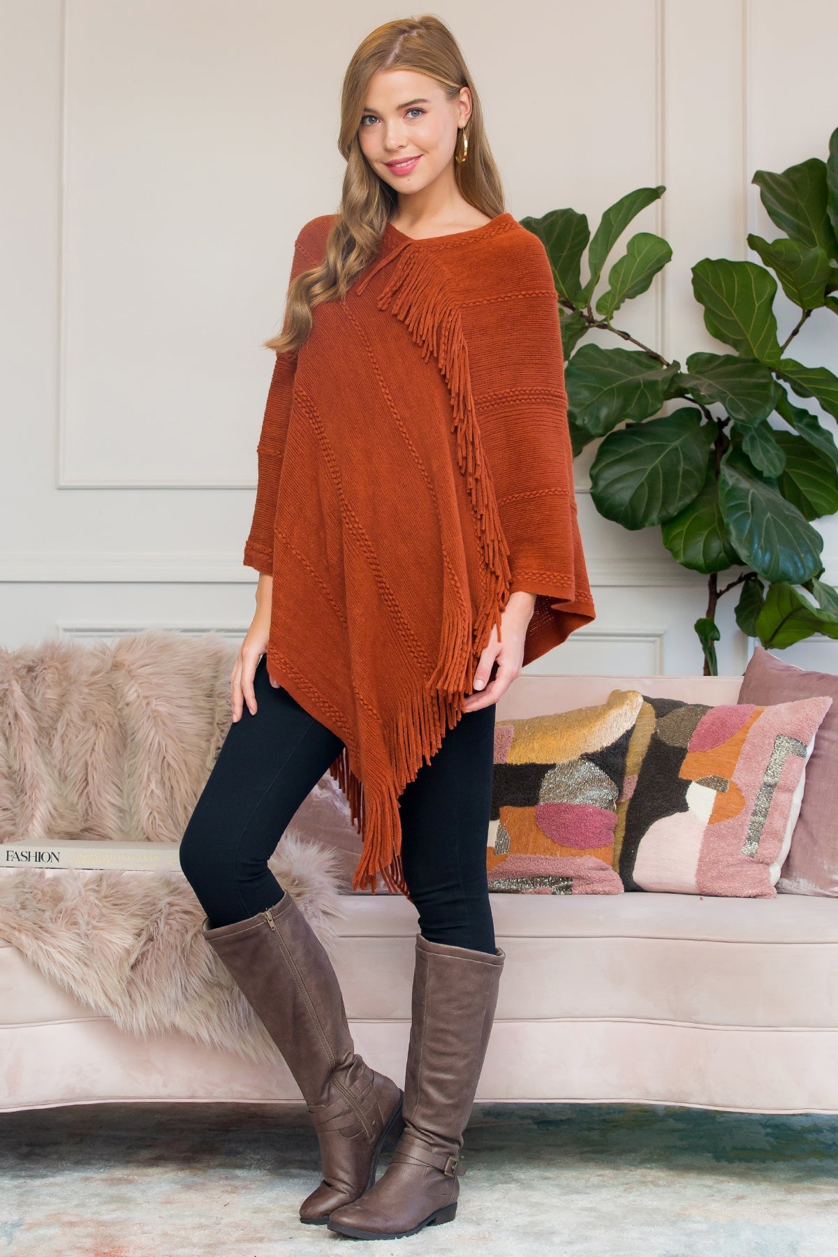 A person stands wearing a Riah Fashion Asymmetric Fringe Poncho in one color, paired with jeans and brown boots, smiling while holding the poncho. In the background are a plant and a sofa with cushions.