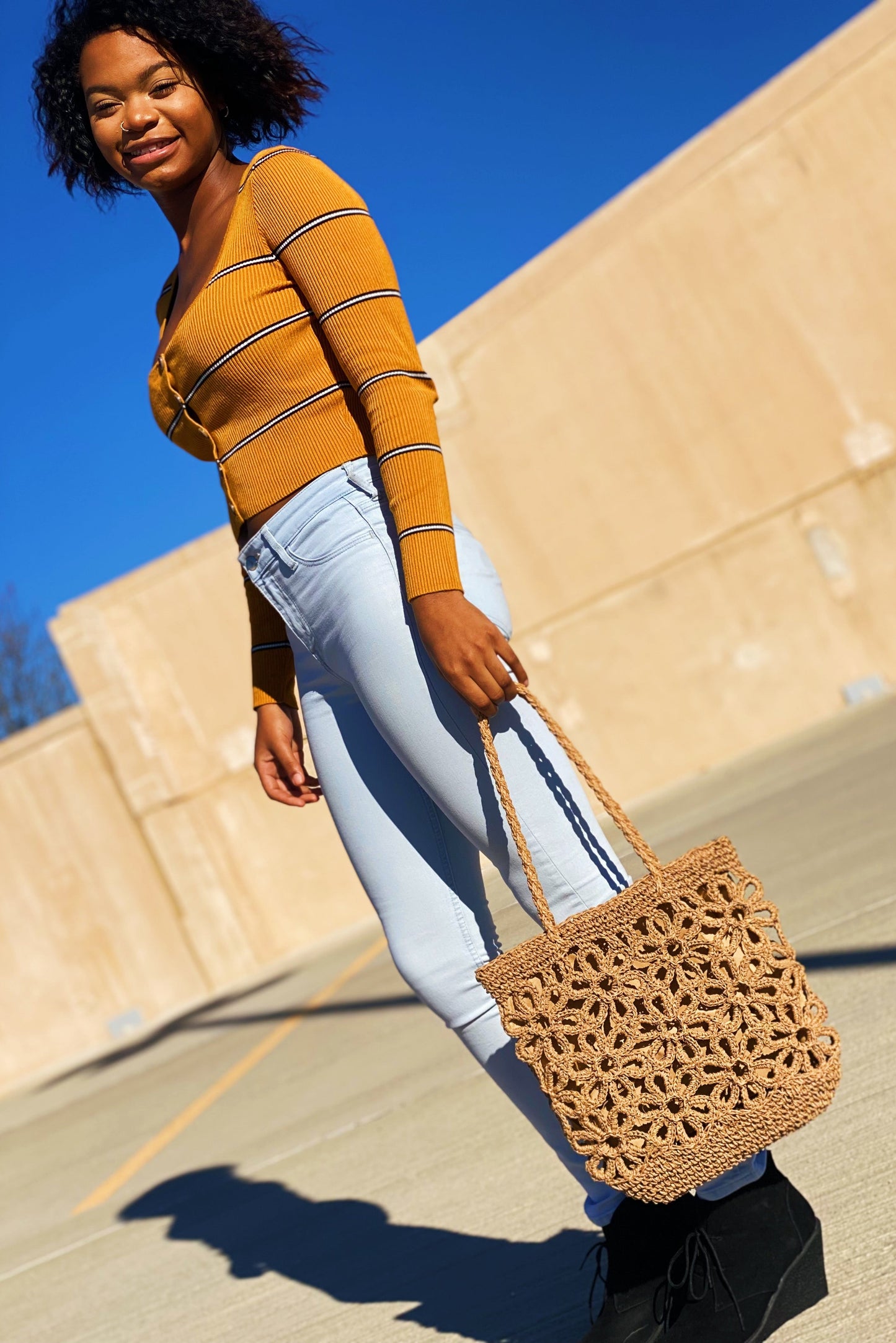 A person smiles while standing outdoors with a clear blue sky and trees in the background, wearing a yellow striped sweater and holding an Ellison + Young Hand Made Muted Floral Tote.