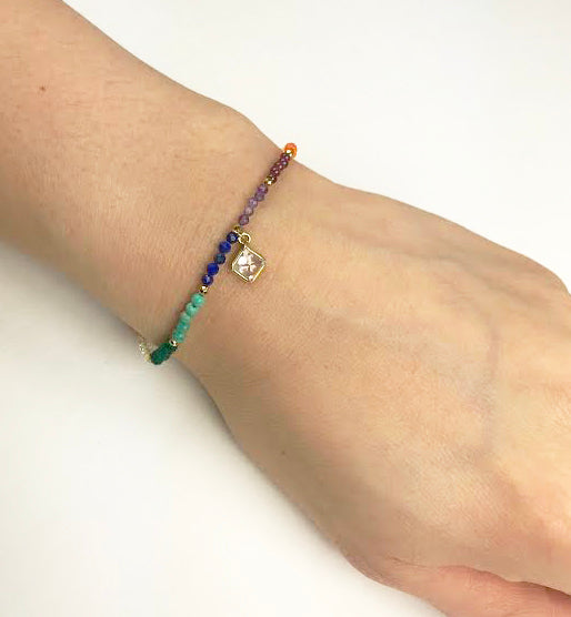 The Chakra With Clear Quartz Drop Bracelet by Gena Myint features colorful beads in green, orange, white, purple, and dark red. It includes a gold clasp, heart charm, and square pendant.