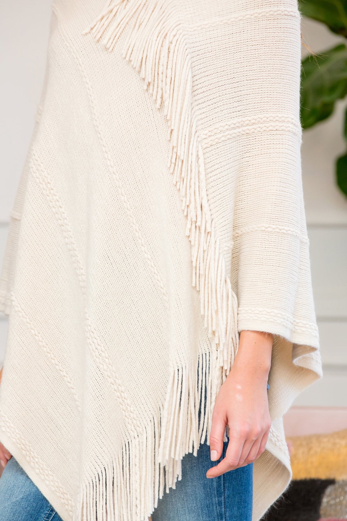 A person stands wearing a Riah Fashion Asymmetric Fringe Poncho in one color, paired with jeans and brown boots, smiling while holding the poncho. In the background are a plant and a sofa with cushions.