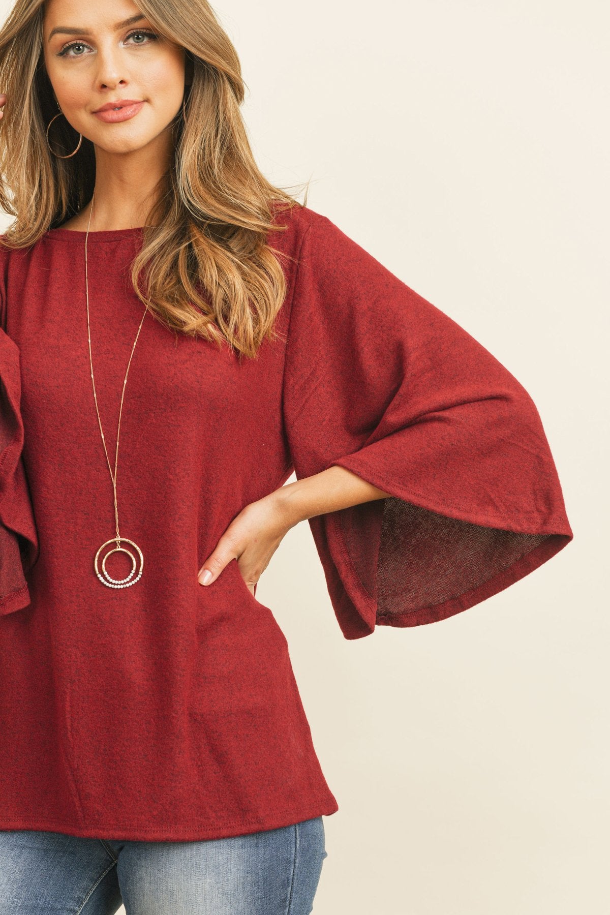 A person in a Riah Fashion Boat Neck Bell Sleeve Solid Hacci Brushed Top and ripped jeans stands against a neutral background, accessorized with hoop earrings and a long necklace.