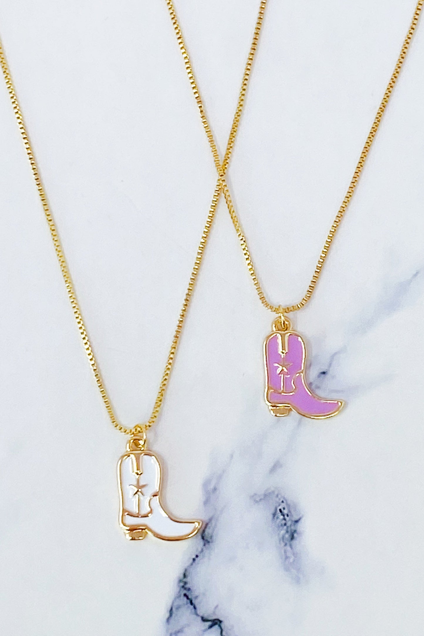 Two Cow Girl Boot Pendant Necklaces by Ellison + Young, one with a white boot and the other with a pink boot, are displayed on a marble surface.