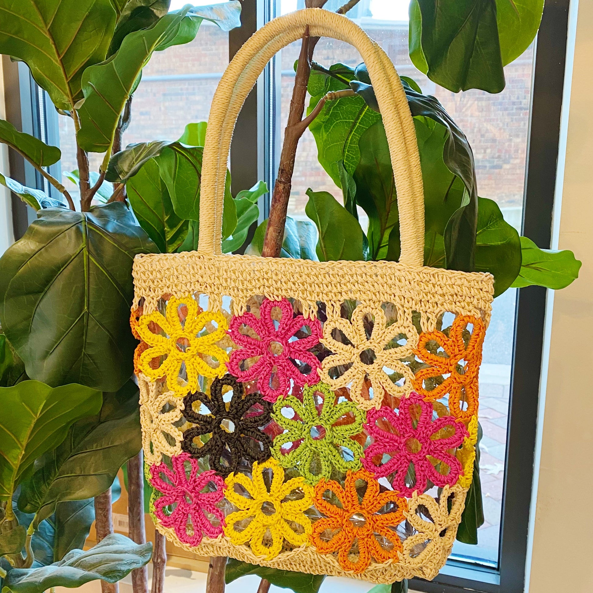 The Hand Made Flower Power Tote by Ellison + Young, featuring colorful floral crochet patterns in orange, green, pink, black, and yellow, rests on dense green foliage.
